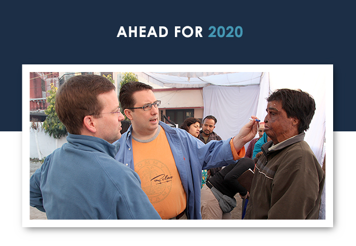 A man in an orange shirt and a man in a blue jacket are seen performing a consult on a burn victim. 
